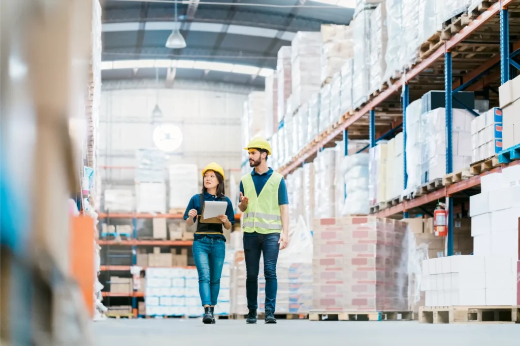 Two workers in a warehouse wearing wearing PPE, holding a clipboard.